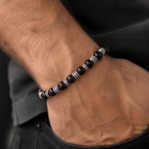 Könnte beinhalten: Ein Perlenarmband mit dunkelbraunen, polierten, kugelförmigen Perlen, die sich mit kleineren, texturierten silberfarbenen Metallzwischenstücken abwechseln. Das Armband besteht aus einer Reihe dieser dunklen Perlen und Metallakzente.
