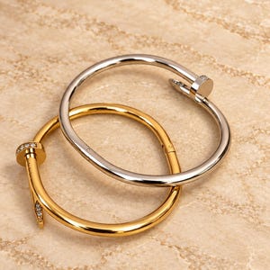 May include: Two circular nail-shaped bracelets, one in polished gold-tone metal and one in polished silver-tone metal. Both bracelets feature a flat circular head and a pointed end, with small clear stones embedded along the head and the tip.