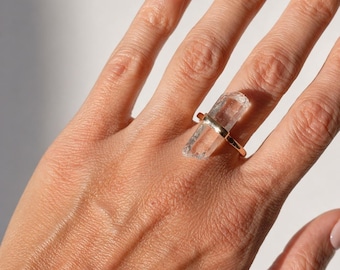 May include: A gold ring with a large, clear crystal. The crystal is rectangular and set horizontally. The ring's design is simple, highlighting the crystal's natural beauty. The ring is a statement piece.