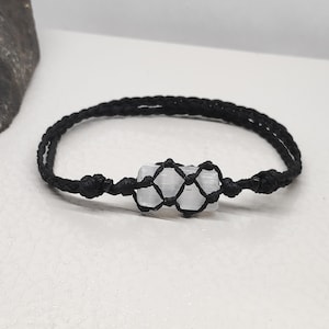 K&ouml;nnte beinhalten: Ein Armband aus schwarzem geflochtenem Band mit einem wei&szlig;en Selenitkristall, der in einen Makramee-Knoten eingewickelt ist.