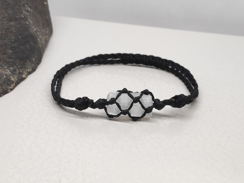 K&ouml;nnte beinhalten: Ein Armband aus schwarzem geflochtenem Band mit einem wei&szlig;en Selenitkristall, der in einen Makramee-Knoten eingewickelt ist.