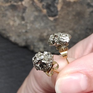 Może przedstawiać: Zbliżenie dwóch kolczyków sztyftów z pirytu. Każdy kolczyk ma skupisko metalicznych, srebrnych kryształów. Kolczyki mają złote sztyfty i zapięcia. Tło jest rozmyte, o neutralnym odcieniu.