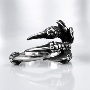 May include: A silver-toned metal ring designed to resemble a bird or dragon claw. The band features a textured, scaly surface that wraps around the finger, ending in three sharp, pointed talons that extend outward from the main circular structure.