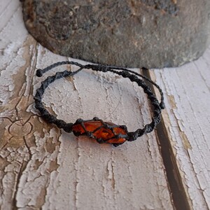 Peut inclure: Un bracelet macramé fait à la main avec une pierre de couleur ambre au centre. Le bracelet est fabriqué avec un cordon macramé noir, présentant un motif tissé. La fermeture réglable assure un ajustement confortable. Le bracelet est présenté sur une surface en bois vieilli.