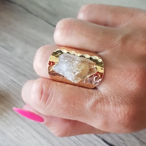 Può includere: Un anello largo color oro caratterizzato da una fascia in metallo martellato e testurizzato. Al centro della fascia si trova un grande cristallo grezzo e traslucido con sfaccettature giallo pallido e trasparenti, dalla forma naturale irregolare.