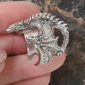 Puede incluir: Un anillo de plata con forma de dragón con alas y escamas detalladas. El anillo está abierto por un lado y se puede ajustar para que se ajuste a diferentes tamaños de dedos.