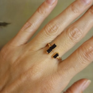 Puede incluir: Un anillo dorado con dos piedras negras rectangulares. El anillo se muestra en una mano, resaltando su diseño minimalista y su estética elegante. Las piedras están dispuestas de forma lineal, añadiendo un toque moderno a la joya.