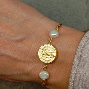 Puede incluir: Un brazalete de cadena dorada con una medalla de San Cristóbal y detalles de perlas blancas.