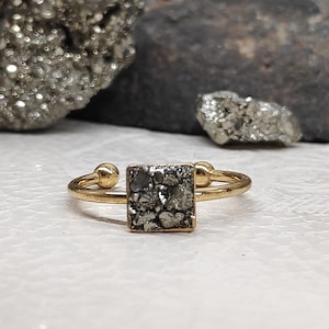Könnte beinhalten: Ein goldener Ring mit einem quadratischen Pyrit-Stein. Der Stein ist dunkelgrau und hat eine raue Textur.