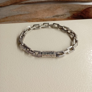 Könnte beinhalten: Ein silbernes Kettenarmband mit einem dekorativen Verschluss. Die Kette besteht aus ineinandergreifenden Gliedern mit aufwendigen Designs.