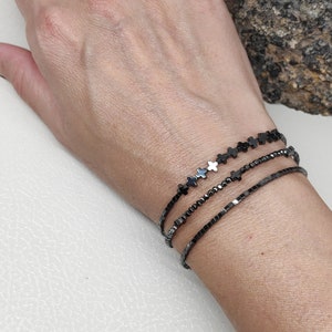 Può includere: Un set di tre braccialetti di perline grigio scuro. Uno presenta perline a forma di croce, un altro ha piccole perline quadrate e il terzo ha minuscole perline rotonde. I braccialetti sono indossati su un polso.