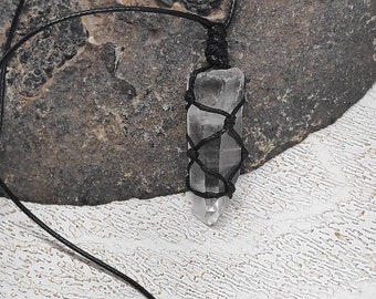 May include: A clear quartz crystal point wrapped in black cord, hanging from a black cord necklace. The crystal is on a gray and white textured surface.