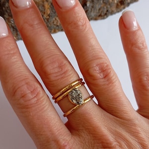 Può includere: Un anello dorato con una fascia strutturata e una pietra a forma di goccia, screziata di grigio. L'anello è indossato su un dito, mettendo in mostra il suo design dettagliato e la finitura metallica. Il design dell'anello è unico e accattivante.