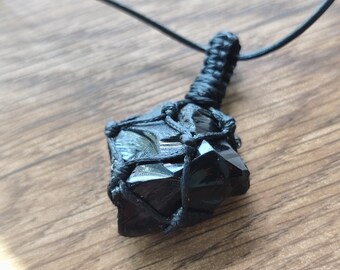 May include: A black obsidian crystal pendant encased in a black macrame cord netting. The raw, glossy stone is held by a woven knot pattern that connects to a thin black cord necklace, creating a secure and functional jewelry piece.