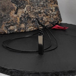 Könnte beinhalten: Eine Halskette aus schwarzem Lederband mit einem schwarzen Metallstab-Anhänger.