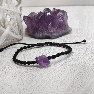 May include: A black cord bracelet with a purple amethyst crystal charm. The bracelet is tied with a knot and has a small black bead at the end.