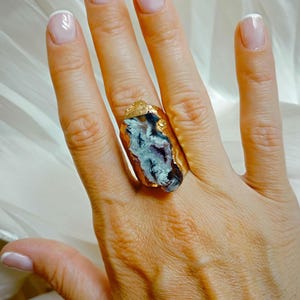Peut inclure: Bague dorée ornée d'une grosse pierre naturelle aux nuances de bleu, violet et blanc. La pierre est sertie dans un anneau doré, créant un bijou unique. Le design de la bague est organique.