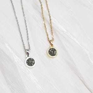 May include: Two delicate necklaces with round pendants. One necklace is silver-toned, the other is gold-toned. Each pendant features a circular, dark, speckled stone. The necklaces rest on a white, textured surface.