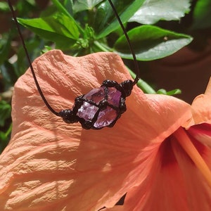 Peut inclure: Un collier en macramé fait à la main avec un cristal d'améthyste brut. Le cristal violet est enfermé dans un filet noir tissé et suspendu à un cordon noir. Le collier repose sur une fleur couleur pêche, avec un feuillage vert en arrière-plan.