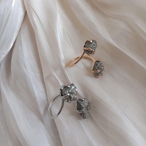 Peut inclure: Deux bagues au design unique. L'une est dorée, l'autre argentée. Chaque bague présente deux éléments rugueux, gris, ressemblant à des pierres. Les bagues sont présentées sur un tissu blanc texturé.