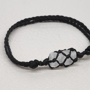 K&ouml;nnte beinhalten: Ein Armband aus schwarzem geflochtenem Band mit einem wei&szlig;en Selenitkristall, der in einen Makramee-Knoten gewickelt ist.