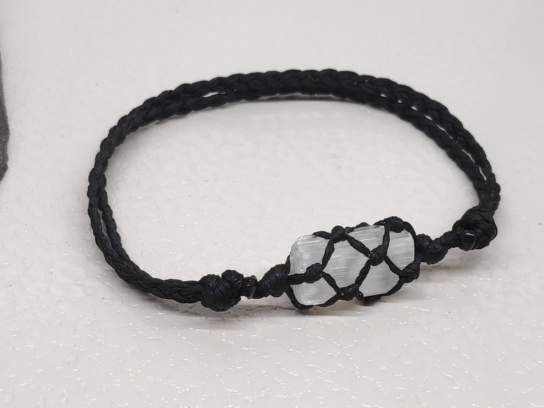 K&ouml;nnte beinhalten: Ein Armband aus schwarzem geflochtenem Band mit einem wei&szlig;en Selenitkristall, der in einen Makramee-Knoten gewickelt ist.