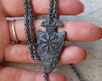 May include: A dark gray metal pendant shaped like an arrowhead with a hammered texture and a black Vegvisir symbol engraved on the front. The pendant hangs from a matching dark gray woven metal chain necklace with a lobster claw clasp closure.