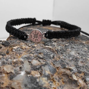 May include: Black braided bracelet with a silver compass charm. The compass charm has the letters N, S, E, and W engraved on it.