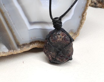 May include: A dark red, raw gemstone pendant necklace. The stone is encased in a black woven net and suspended from a black cord. The background is a white surface with a gray and white patterned stone.
