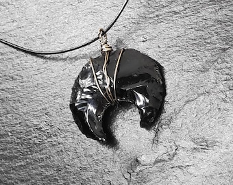 May include: A black obsidian crescent moon pendant wrapped in gold wire, hanging from a black leather cord.