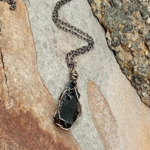 May include: A black obsidian pendant wrapped in copper wire, hanging from a silver chain necklace. The pendant is a rough, natural shape and is set against a rustic wooden background.