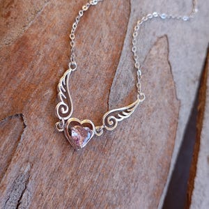 Op de afbeelding: Een zilveren ketting met een hartvormige hanger met een roze edelsteen. Het hart wordt omringd door twee zilveren vleugels.