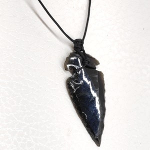 May include: A black obsidian arrowhead pendant on a black cord necklace. The arrowhead is a dark, iridescent black with a sharp point.