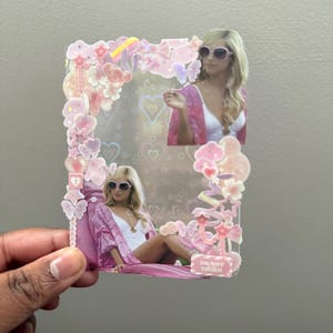 Könnte beinhalten: Ein klarer, rechteckiger Aufkleber mit einem rosa Blumen- und Herzrand. Der Aufkleber zeigt zwei Bilder einer Person mit Sonnenbrille und einem rosa Bademantel. Der Text "DO NOT TOUCH" ist sichtbar.