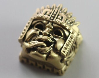 Könnte beinhalten: Eine detaillierte, goldfarbene Skulptur eines stilisierten Gesichts mit einer Krone. Das Gesicht hat einen markanten Schnurrbart und einen strengen Ausdruck. Die Skulptur ist quadratisch und scheint aus Metall zu sein.