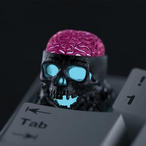 May include: A black skull keycap with a glowing blue light inside and a pink brain on top. The keycap is placed on a keyboard with the "Tab" key visible.