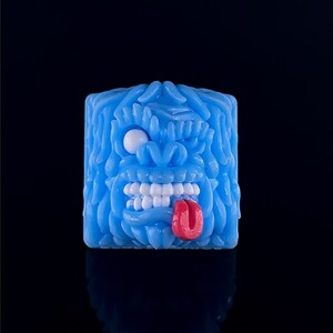 Könnte beinhalten: Ein blauer und weißer Keycap mit einem detaillierten, strukturierten Design eines Monstergesichts mit einem weißen Auge, weißen Zähnen und einer roten Zunge, die herausragt.
