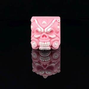 Puede incluir: Un keycap rosa y blanco con un diseño de calavera. La calavera tiene un diseño detallado con líneas y patrones intrincados.