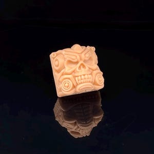 Puede incluir: Objeto decorativo cuadrado de color beige claro con un diseño de calavera tallada. La calavera tiene detalles, incluyendo cuencas de los ojos y dientes. El objeto se refleja en una superficie negra brillante.