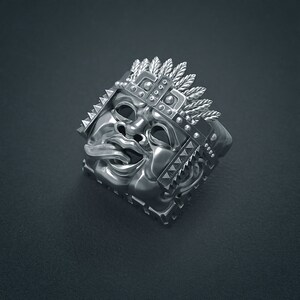 K&ouml;nnte beinhalten: Ein silberner Ring mit einem detaillierten Design eines Gesichts mit herausgestreckter Zunge. Das Gesicht ist von einer Krone aus Federn und Stacheln umgeben.