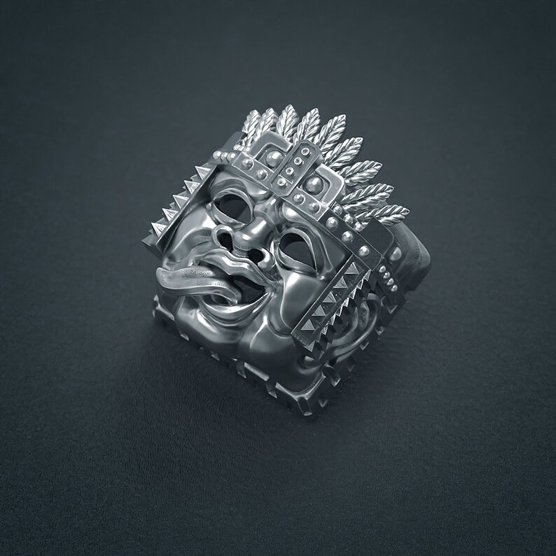 K&ouml;nnte beinhalten: Ein silberner Ring mit einem detaillierten Design eines Gesichts mit herausgestreckter Zunge. Das Gesicht ist von einer Krone aus Federn und Stacheln umgeben.