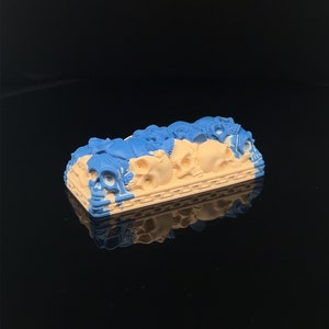 Puede incluir: Un keycap impreso en 3D azul y beige con un diseño de calavera. El keycap está hecho de resina y tiene un acabado liso.