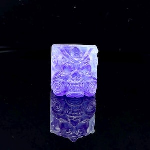 Puede incluir: Un keycap de resina morado y blanco con un diseño de calavera detallado. El keycap es translúcido y tiene un acabado brillante.