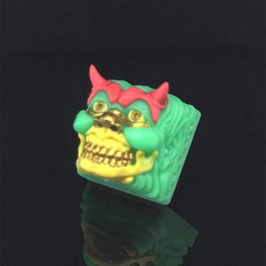 May include: A green and red keycap with a detailed skull design and gold accents. The keycap is shaped like a mythical creature with horns and a fierce expression.