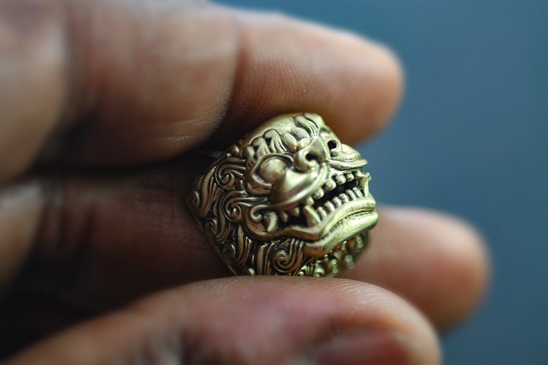 May include: A gold-colored ring with a detailed design of a mythical creature's face. The creature has a wide mouth with sharp teeth and a prominent nose.
