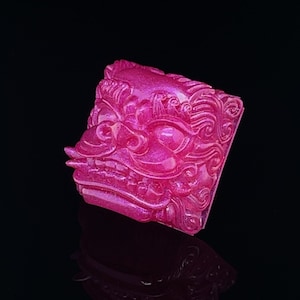 May include: A pink resin keycap with a detailed, intricate design of a mythical creature's face. The keycap has a glossy finish and a textured surface.