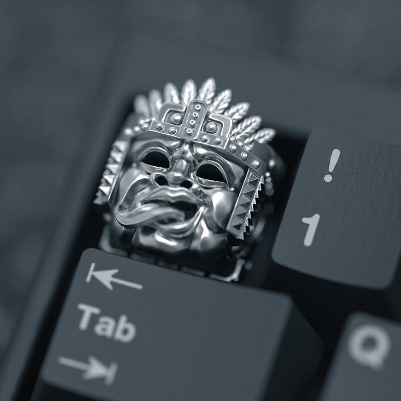 K&ouml;nnte beinhalten: Eine silberfarbene Tastaturkappe mit einem detaillierten Design einer Maya-Maske mit Federkopfschmuck. Die Tastaturkappe liegt auf einer schwarzen Tastatur.