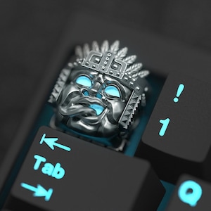 K&ouml;nnte beinhalten: Eine silberne Tastenkappe mit einem detaillierten Design eines Gesichts, das eine Kopfbedeckung tr&auml;gt. Die Tastenkappe ist mit blauem Licht beleuchtet und befindet sich auf einer schwarzen Tastatur.