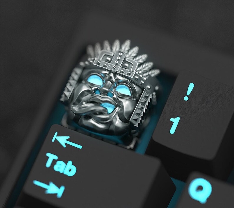 K&ouml;nnte beinhalten: Eine silberne Tastenkappe mit einem detaillierten Design eines Gesichts, das eine Kopfbedeckung tr&auml;gt. Die Tastenkappe ist mit blauem Licht beleuchtet und befindet sich auf einer schwarzen Tastatur.