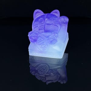 Puede incluir: Una figurita de resina de Maneki-Neko, un gato de la suerte japonés, con su pata derecha levantada. La figurita está en posición sentada con una amplia sonrisa y una moneda en su pata. La figurita es de color morado y blanco con un degradado.